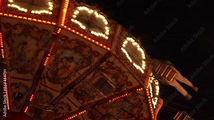 close-up night view of an ornate carousel in motion, illuminated by bright lights against a dark sky. The carousel's intricate designs and vibrant colors are highlighted by rows of lights that create