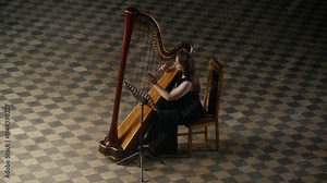 High angle full shot of skillful young female Caucasian harpist in fancy black gown with long loose hair performing solo on harp with dignity and inspiration in hall with checkerboard floor Stock Video
