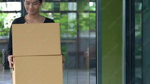 4K Wide shot of adult asian businesswoman holding boxes walking through office building. Office woman being fired carrying her belongings boxes and walking outside the office building.