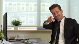 An office worker in a suit sits at a desk in front of a computer, points a finger at the camera and nods with a smile