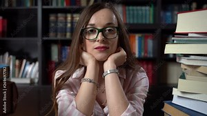 Beautiful young librarian woman in glasses sitting on bookshelf background in school library
