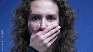 Scared woman reacts with shock and unbelief by covering mouth feeling stress and preoccupation, looking directly at camera with blue background. Close-up face 20s person Stock Video