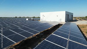 Solar Farm with Battery Storage System - An aerial view of a large solar farm on a sunny day. Rows of solar panels extend across the landscape, with a modern white building housing the battery