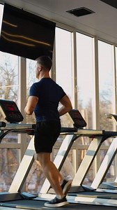 Athletic man running on a treadmill in a modern fitness center, with morning sunlight streaming through large windows. Cardio and healthy lifestyle concept