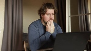 Scared man at laptop, experiencing fear and anxiety while typing, working remotely in a home office setting.