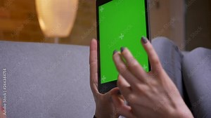 Closeup back view shoot of female hands holding the tablet vertically with green screen lying on the couch indoors in a cozy apartment