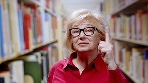 Caucasian blonde woman old teacher working in the library wearing glasses pointing up finger telling to be queit keeping order. Real portrait.