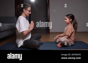 Mother and daughter sitting in lotus pose with namaste hands, smiling, and finding calm performing mindful meditation and wellness exercises together in a home setting