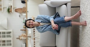 Vertical view optimistic middle-aged woman smile looks at camera relaxing alone on sofa in studio apartment. Portrait of happy carefree untroubled retired female, rest, enjoy weekend leisure at home