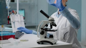 Close up of microscope tool while scientist analyzing petri dish and using modern computer in background. Woman at laboratory desk with blood sample and tray of vacutainers for examination
