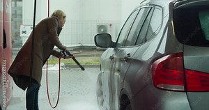 Cheerful young blonde washes her car in a self-service car wash. Cheerful young blonde washes her car in a self-service car wash. Beautiful business lady washes her car with a high-pressure water jet