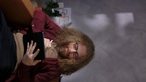 Young Happy Man with Long Curly Ginger Hair and Wearing Glasses Working Remote from Home on his Digital Tablet sitting on Sofa at Home, Freelance Designer Working, Vertical Video Format, Copy Space