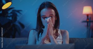 Sad young woman worrying while watching an emotional scene in movie. Upset girl with laptop on her knees looking at display, crying and using paper tissue. Home background.