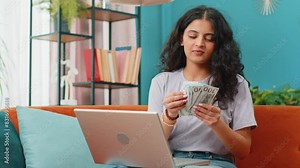 Planning budget. Rich happy Indian woman counting money cash use laptop computer calculate domestic bills at home. Arabian girl satisfied of income earnings, saves money for planned vacation, gifts