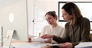 Attentive women look at monitor and investigation papers comparing statistics. Female colleagues sit at table near computer in office slow motion