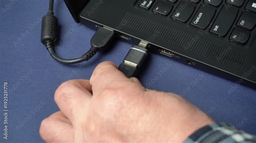 Hand inserting USB connector into laptop port, showing progressive action of connecting device to computer with visible cables and workspace