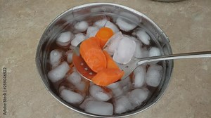 Home cooking - Checking or string with skimmer blanched or precooked slices of carrots being cooling down in ice bath in stainless steel bowl.