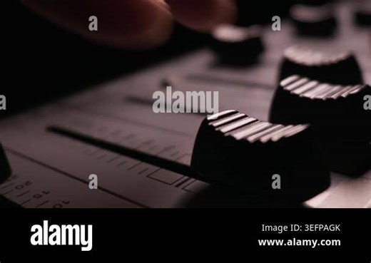 A finger pulls down a fader knob on an audio mixing console to decrease the volume. Close-up details highlight the ribbed plastic sliders. Dark studio lighting surrounds the soundboard Stock Video Footage - Alamy