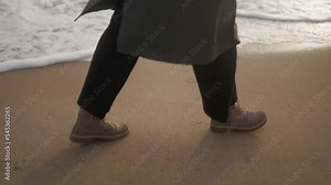 Side view of female feet in boots walking along sea waves foam on sandy beach. Woman in green coat goes along ocean tides on coastline. Tilting establishing dreamy cinematic depersonalized shot.