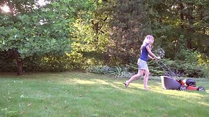 Woman working in garden cutting grass with lawn mower. Evening sunlight penetrate through tree branches. Static shot. 4K