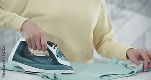 A woman in a yellow sweater holds an iron and irons clothes. The iron releases steam. Medium shot, face is not visible.4K Stock Video