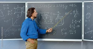 Caucasian good-looking male teacher standing at blackboard with formulas, graphics and laws and explaining with pointer. Lecturer of mathematics at school or college. Math lection.