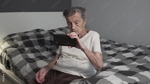 Senior woman in her 90s learns how to use a smartphone sitting on her bed at home. Elderly female gets educated on how to use phone in retirement home. Old lady studies using technology.
