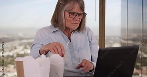 Senior professional woman on lunch break eating and using laptop in office, Caucasian lady eating Chinese take-out while looking at portable laptop in office setting, 4k