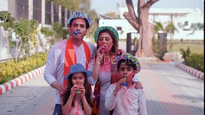 A happy Indian family with their faces smeared in Holi colors blowing blow-out whistles - Holi celebrations childhood games leisure time. A young couple and little kids celebrating Holi in a publ...
