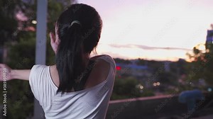 Rear view of Young active Asian female stretch her arms and shoulder watching sunset and city traffic jams from apartment balcony, flexible upper body part, beautiful and relax, exercise motivation