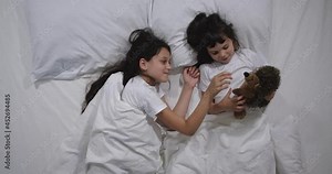 Top view of Caucasian two Children fighting for a toy in bed with white sheets. High angle shooting. Little sisters argue, take offense at each other and turn away, then they make up and play