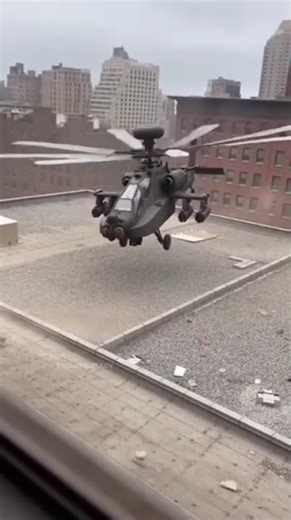 Apache attack helicopter hovering low over a city building rooftop, kicking up dust.