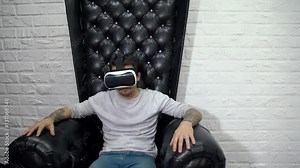A man watches 360 degree video virtual reality using wearable head mounted display, sitting in a black large leather armchair.