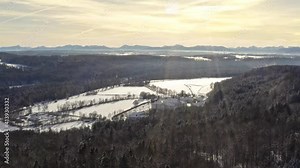 Yellow glowing sky over the alp mountains in a winter landscape, flying down by drone to hide the view behind some trees of a forest.