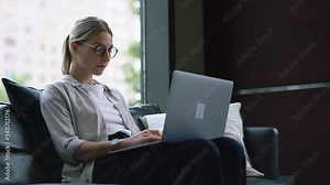 Young woman working remotely on internet content at laptop computer while got paper documents with financial reports in modern open space office.Hipster girl installing app on netbook