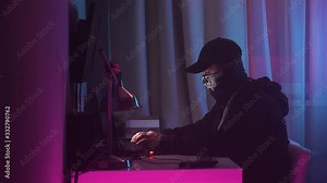 Male hacker sits at his desk and breaks into the system. Man in black hoodie with bandana and black cap hides his face and works at laptop at neon lighting. Police siren background. Smoke in room.