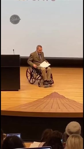 A man in a wheelchair rolls onto the stage for what seems like a normal lecture. In front of him lies a wooden floor with a strange, pointed structure in the center. Without hesitation, he drives straight over it while the audience watches. Suddenly, the middle section collapses and the floor breaks open. The wheelchair and the man drop through the hole, shocking the crowd. Papers fly into the air as he disappears into the opening. The audience is left stunned, unsure if it was an accident or a 