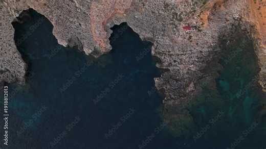 Top-down aerial of southern Malta coast showing calm turquoise waters, a wrecked ship, rocky formations, and garrigue vegetation. Drone moves forward slowly revealing contours, trails, and cliffs.