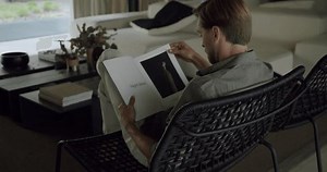 Back view on young man sitting on comfort chair, flipping and reading magazine while resting in lounge room at modern apartment. Male with book relaxation at home at leisure time