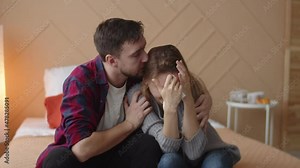Man calms crying woman sitting on bed in bedroom, hugs and kisses her, front view. Couple, man tries to comfort crying wife. Sad couple comforting each other sitting on a couch in the living room at