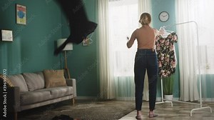 Girl in jeans and brown sweater in front of window removes dress from hanger and scatters it around spacious room