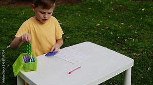 Child with autism has math therapy lesson: uses colorful Numicon shapes, counts, writes answers on paper; sits at white table in sunny garden with bright green grass
