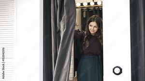 Young woman is standing in a changing room in a shop. She is pulling the curtain to demonstrate her choice, smiling and pulling it back. Locked down real time medium shot