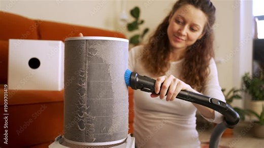 Diligent woman using a vacuum cleaner with a brush attachment to carefully remove a thick layer of dust and allergens from a cylindrical air purifier filter, improving indoor air quality