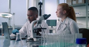 Advanced medical science laboratory: Two diverse scientists using laptop to develop new drug formula or vaccine, doing microscope analysis of samples from test tubes. Medicine and pharmaceuticals.
