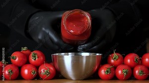 Vegetarian Ketchup pouring from a glass bottle in sauceboat over black background, fresh tomatoes, garlic, pepper and spice near. Adding natural red sauce from bottle. Slow motion, close-up, macro