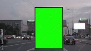 Modern billboard with a green screen on a busy defocused highway with traffic, neon lights, city buildings at background. Blank billboard for advertising.