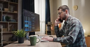 Focused man sitting in front of computer screen. Male talking by phone and making notes with pencil. Man communicating by smartphone and looking at gadget screen. Male working with cryptocurrency.