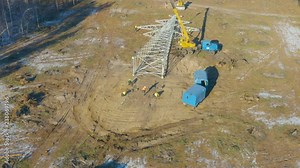 Aerial circling view of the new power line pylon (transmission tower or power tower) lying on the ground and lot of steel erector workers preparing it for installation