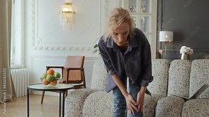 Woman in 60s sitting on couch in living room with laptop. She works or enjoys leisure online activities. tries to stand up and feels unexpected sharp knee pain causes her to frown and hold her knee